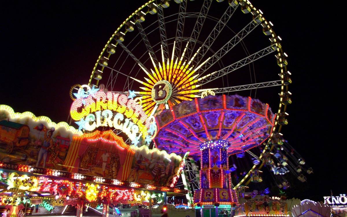 Ein beleuchtetes Riesenrad vor dunklem Nachthimmel. Davor weitere bunte Karussells.