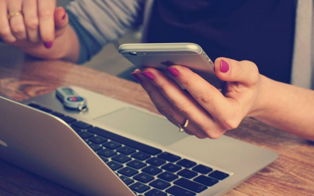 Eine Person sitzt am Laptop. Währenddessen hält sie in ihrer anderen Hand ein Smartphone.