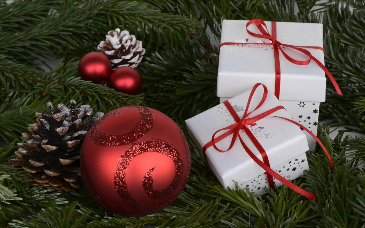 Zwei kleine Geschenkpäckchen in weißem Papier und rotem Geschenkband auf Tannengrün. Dazu zwei rote Weihnachtskugeln und Tannenzapfen.