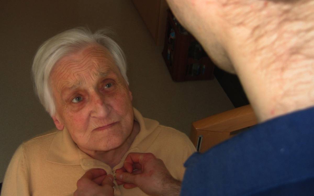 Eine Seniorin, der eine andere Person die Bluse zuknöpft, die man nur von hinten sieht (Kopf, Nacken, Schulter)