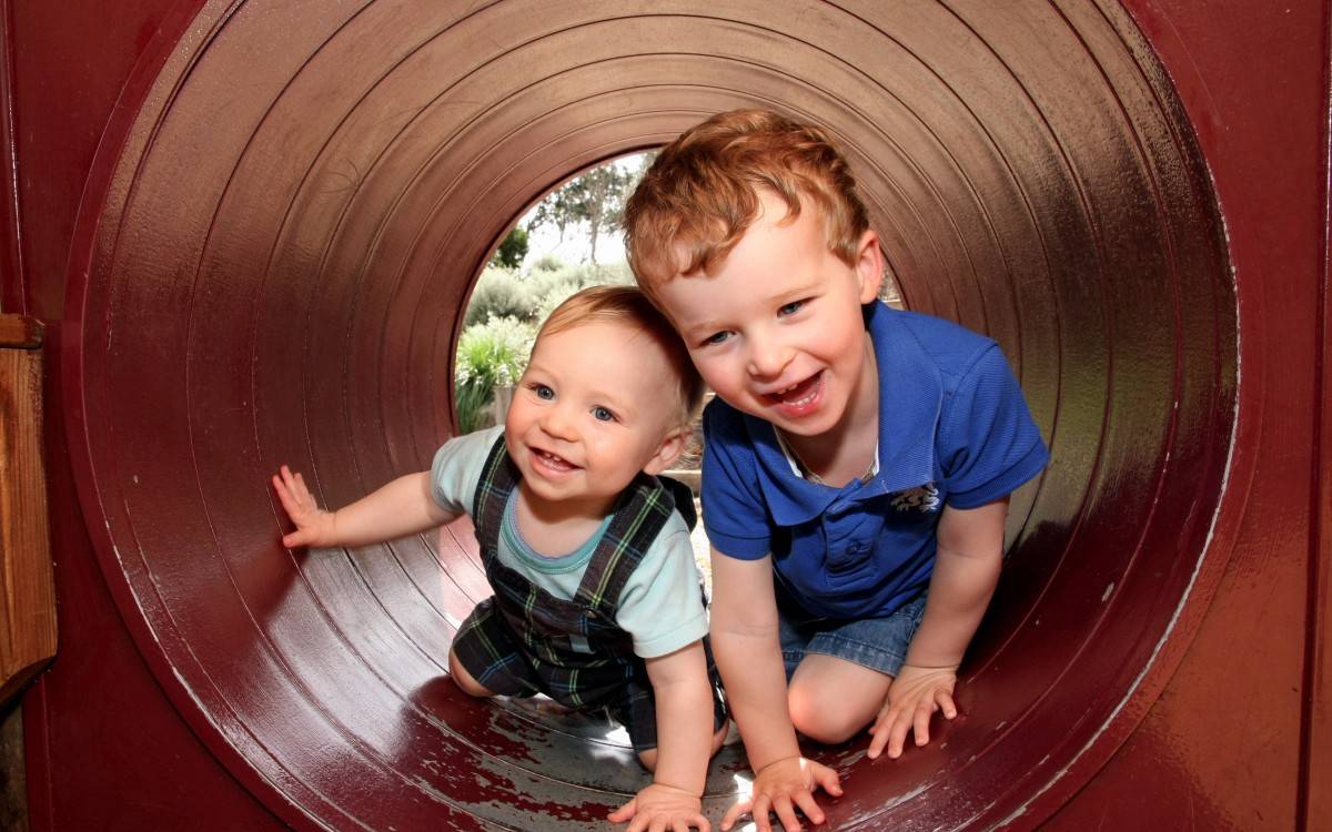 Zwei kleine Jungen lachend in einer Röhre, offenbar auf einem Spielplatz. Durch die Röhre sieht man im Hintergrund grüne Natur und einen Baum.