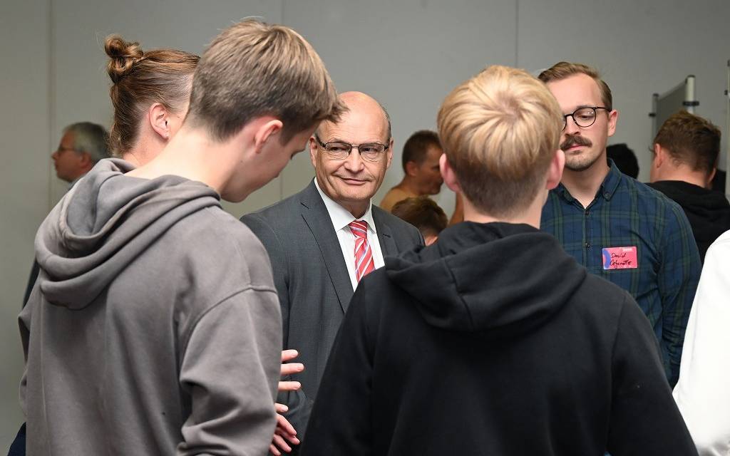 Menschengruppe im Gespräch: Zwei Jugendliche von hinten. Im Zentrum lächelt der Landrat.