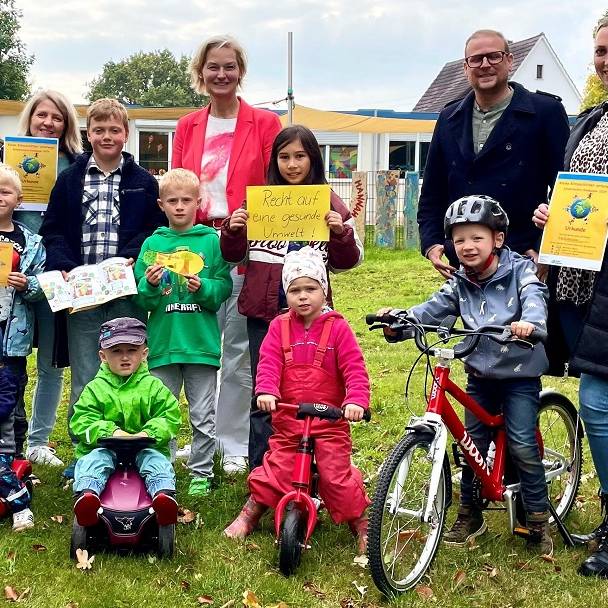Kindergarten- und Schulkinder in zwei Reihen auf einer Rasenfläche. Vorne die Kleinen mit Bobbycars, Rollern und Fahrrädern, dahinter die Größeren mit Umweltschutz-Transparenten. Ganz hinten Erwachsene.