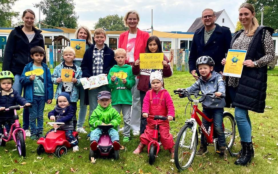 Kindergarten- und Schulkinder in zwei Reihen auf einer Rasenfläche. Vorne die Kleinen mit Bobbycars, Rollern und Fahrrädern, dahinter die Größeren mit Umweltschutz-Transparenten. Ganz hinten Erwachsene.