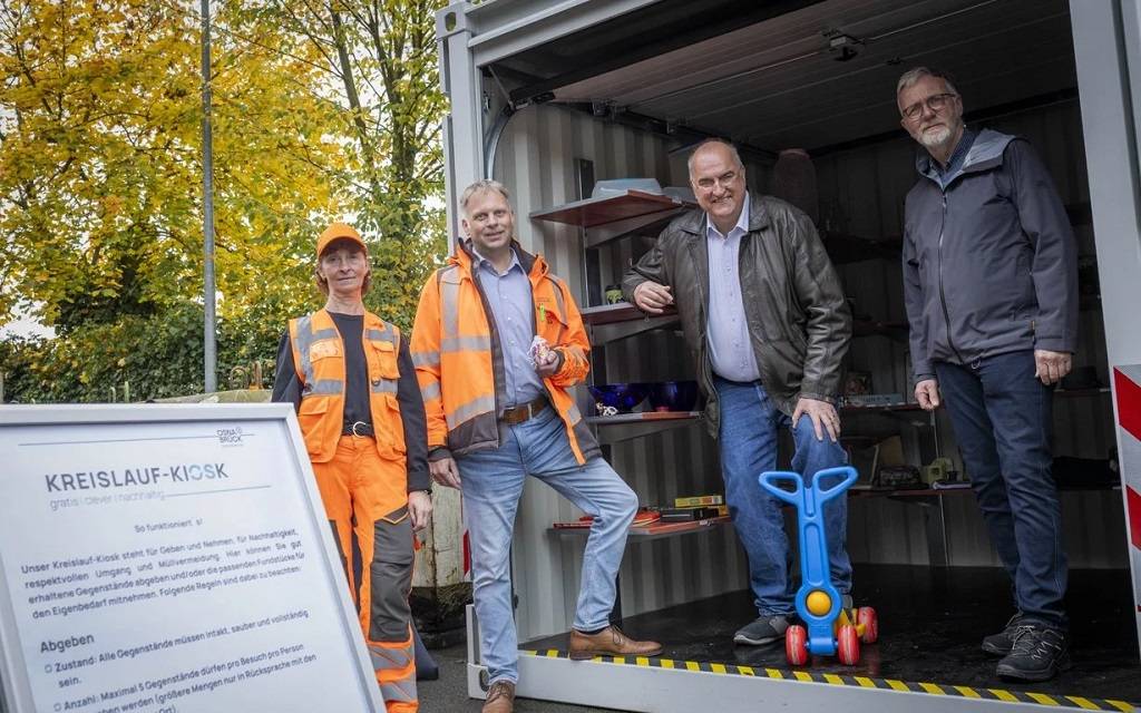 Ein Container mit Regalen voller weiterverwertbarer Dinge. Im Vordergrund eine Frau und ein Mann in orangener Arbeitskleidung und zwei weitere Männer bei der Eröffnung des Kreislauf Kiosk
