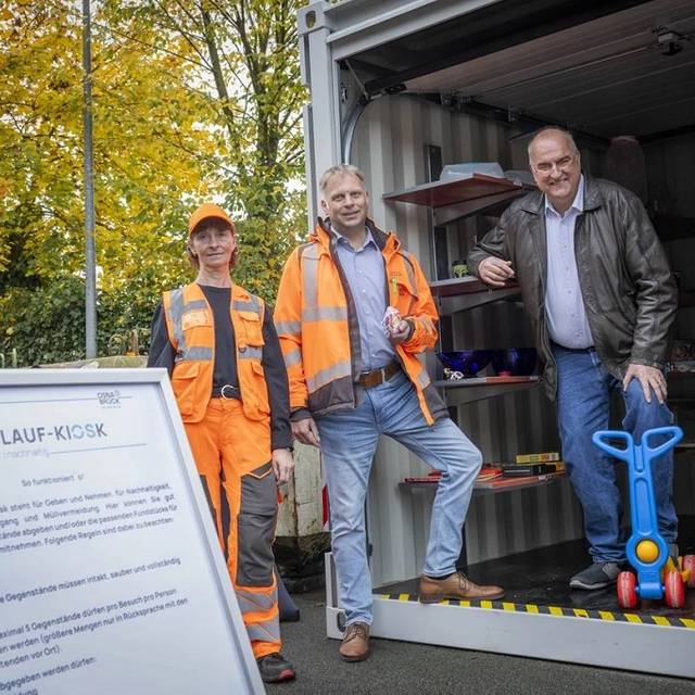 Ein Container mit Regalen voller weiterverwertbarer Dinge. Im Vordergrund eine Frau und ein Mann in orangener Arbeitskleidung und zwei weitere Männer bei der Eröffnung des Kreislauf Kiosk