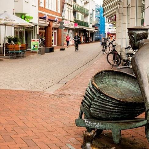Eine Fußgängerzone mit Außengastronomie und Gästen. Im Vordergrund eine Bronzestatue zur Regionalgeschichte