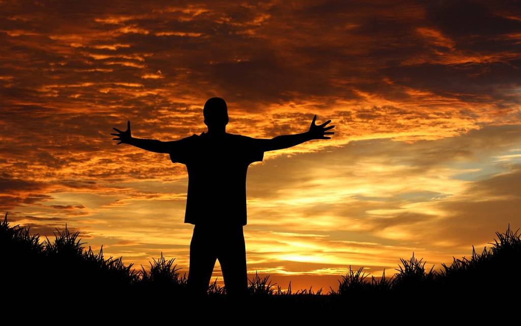 Junger Mann - von hinten aufgenommen, backlighted - breitet die Arme aus vor sonnenrotem Himmel