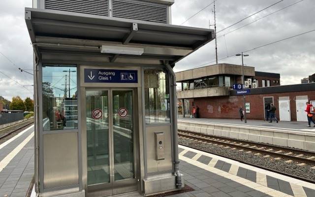 Ein moderner barrierefreier Bahnsteig mit Blindenleitsystem und Aufzug im Vordergrund. Im Hintergrund das ibbenbürener Bahnhofsgebäude