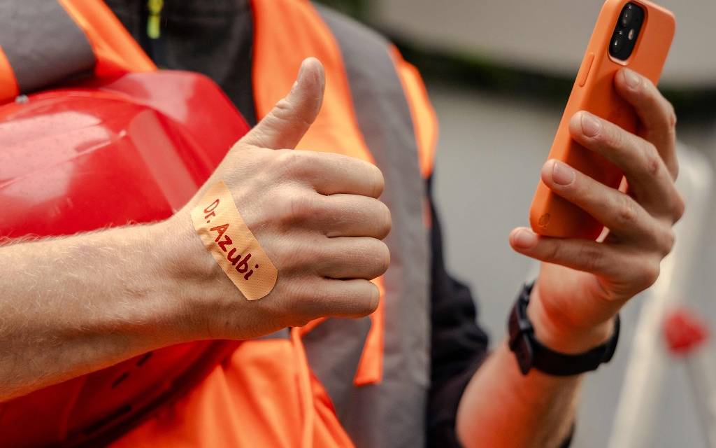 Brustausschnitt eines Arbeiters in orangener Signalkleidung. In der linken Hand ein orangenes Handy, auf dem rechten Handrücken ein orangenes Pflaster mit der Aufschrift "Dr. Azubi"