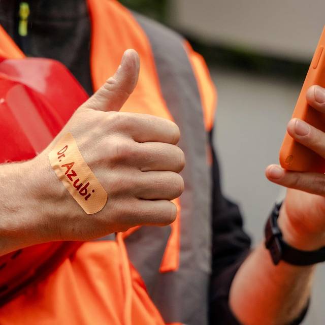 Brustausschnitt eines Arbeiters in orangener Signalkleidung. In der linken Hand ein orangenes Handy, auf dem rechten Handrücken ein orangenes Pflaster mit der Aufschrift "Dr. Azubi"
