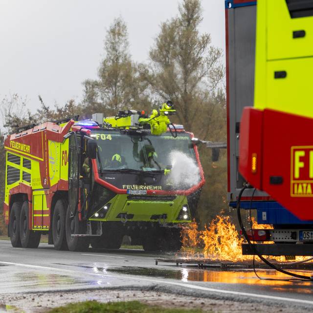 Ein Feuerwehrauto fährt über eine Straße am Flughafen Münster/Osnabrück. Vor dem Auto brennt es.