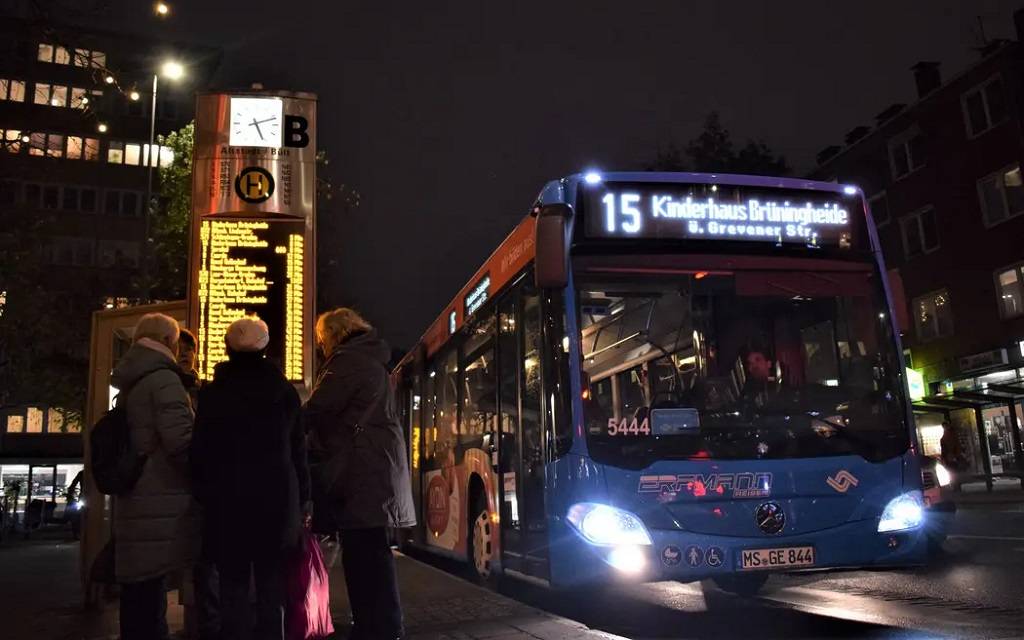 Ein Linienbus im Dunklen und Menschen, die an der Bushaltestelle stehen. Im Hintergrund ein digitaler Fahrplan.