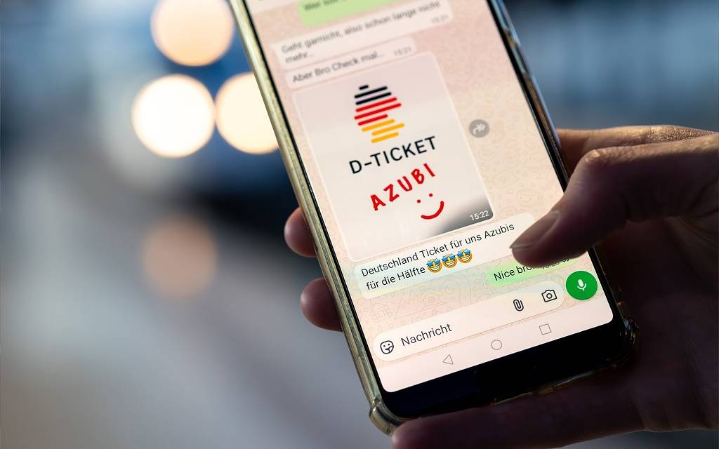 Ein Handy in einer Hand. Auf dem Bildschirm eine WhatsApp-Meldung mit Bild: D-Ticket AZUBI und ein Smiley. Darunter eine Nachricht: Deutschland Ticket für uns Azubis für die Hälfte. Und drei Strahle-Smileys
