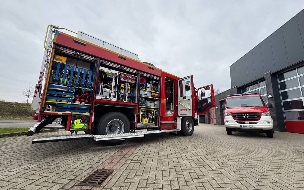 Zwei Feuerwehrfahrzeuge - eins davon offen und voller Technik - auf einem gepflasterten Platz vor einer Halle mit Toren.