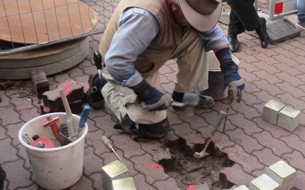 Mann mit Hut in Arbeitskleidung und Weste kniet vor aufgenommenem Straßenpflaster. Stolpersteine liegen bereit, um sie in das Loch einzusetzen.