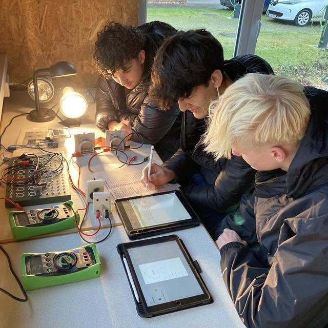 Drei Oberstufenschüler an einem Tisch voller Messegräte, Technik und Tablets. einer füllt eine Tabelle aus