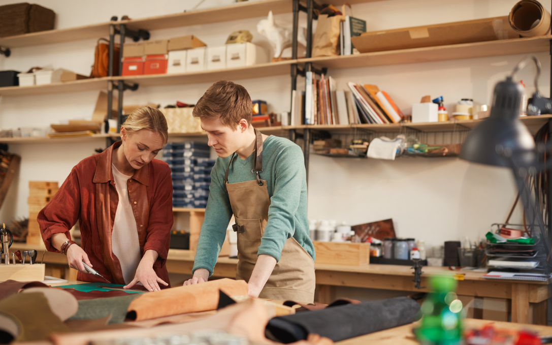 Junge Frau und junger Mann in einem Werkstattumfeld an einem Tisch beim Leder zuschneiden. Sie hat ein Cuttermesser in der Hand, er schaut ihr zu.