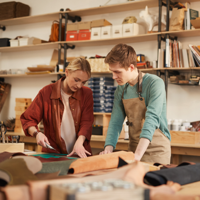 Junge Frau und junger Mann in einem Werkstattumfeld an einem Tisch beim Leder zuschneiden. Sie hat ein Cuttermesser in der Hand, er schaut ihr zu.