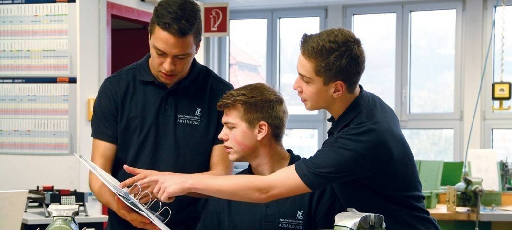 Drei junge Azubis in einer Lehrwerkstatt diskutieren über Daten in einer Mappe, die einer von ihnen hält