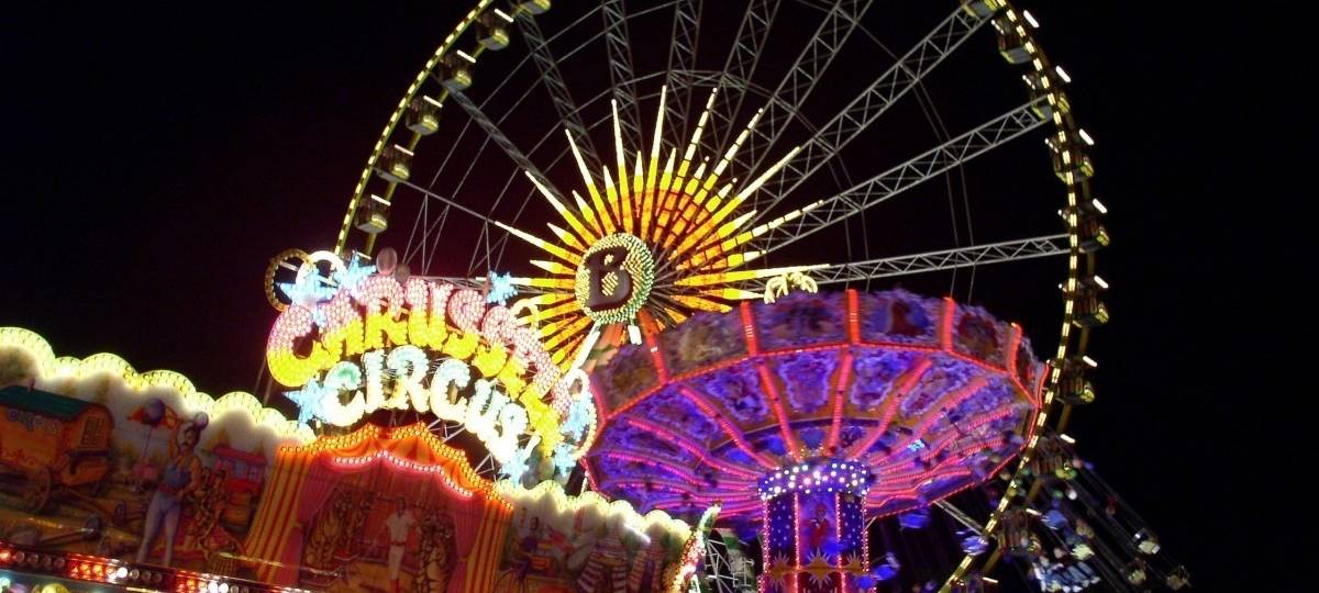 Ein beleuchtetes Riesenrad vor dunklem Nachthimmel. Davor weitere bunte Karussells.