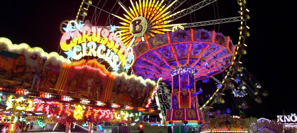 Ein beleuchtetes Riesenrad vor dunklem Nachthimmel. Davor weitere bunte Karussells.