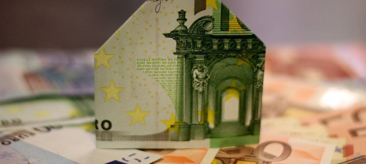 Ein Haus aus einem 100-Euro-Schein gefaltet steht auf weiteren Geldscheinen
