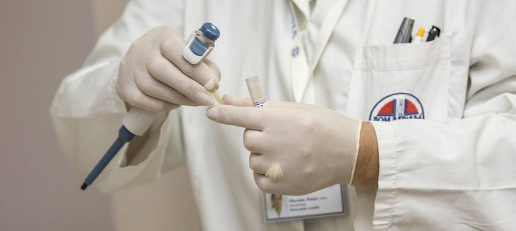 Ärztin oder Arzt (Kopf sieht man nicht) im weißen Kittel mit OP-Handschuhen, Pipette und Ampulle in der Hand.