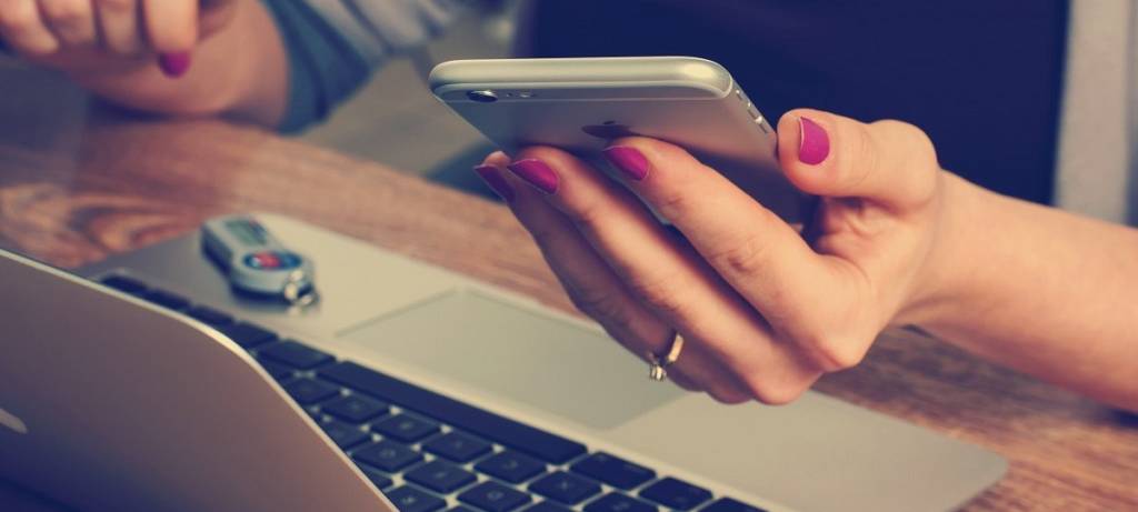 Eine Person sitzt am Laptop. Währenddessen hält sie in ihrer anderen Hand ein Smartphone.