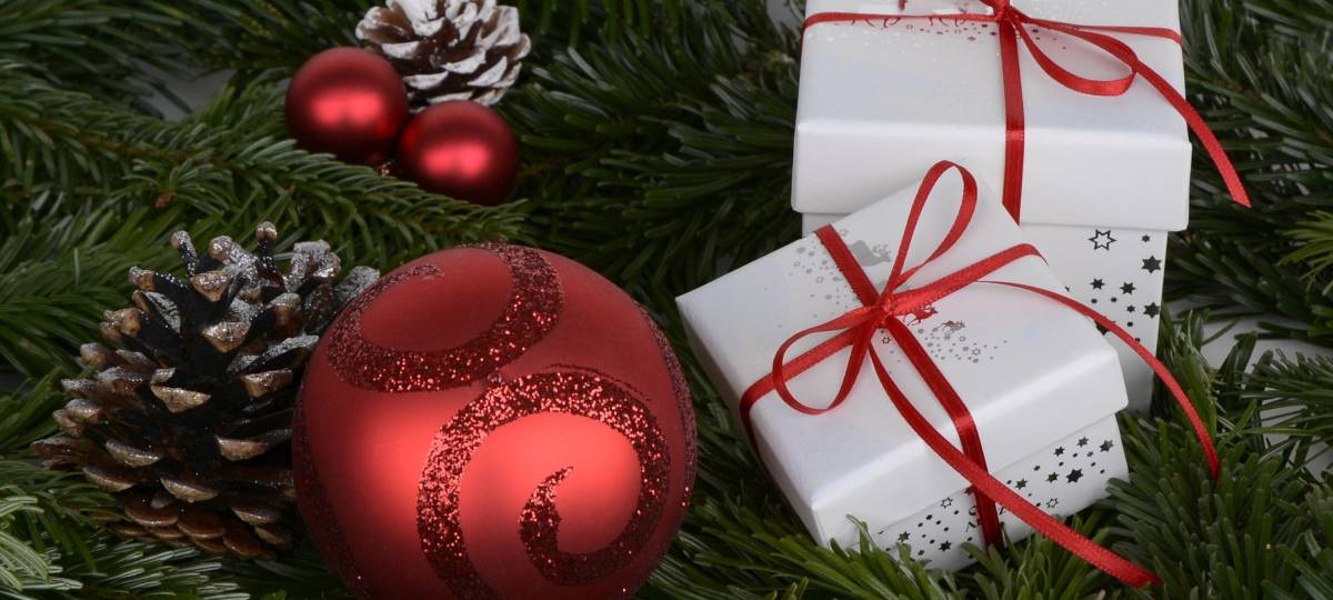 Zwei kleine Geschenkpäckchen in weißem Papier und rotem Geschenkband auf Tannengrün. Dazu zwei rote Weihnachtskugeln und Tannenzapfen.