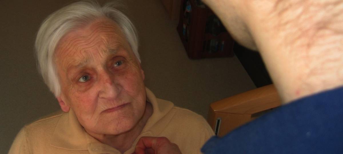 Eine Seniorin, der eine andere Person die Bluse zuknöpft, die man nur von hinten sieht (Kopf, Nacken, Schulter)