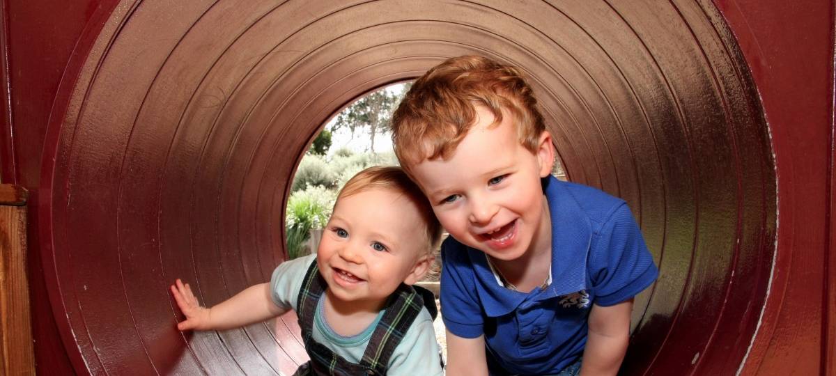 Zwei kleine Jungen lachend in einer Röhre, offenbar auf einem Spielplatz. Durch die Röhre sieht man im Hintergrund grüne Natur und einen Baum.