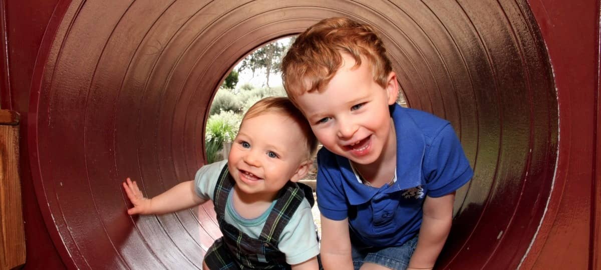 Zwei kleine Jungen lachend in einer Röhre, offenbar auf einem Spielplatz. Durch die Röhre sieht man im Hintergrund grüne Natur und einen Baum.