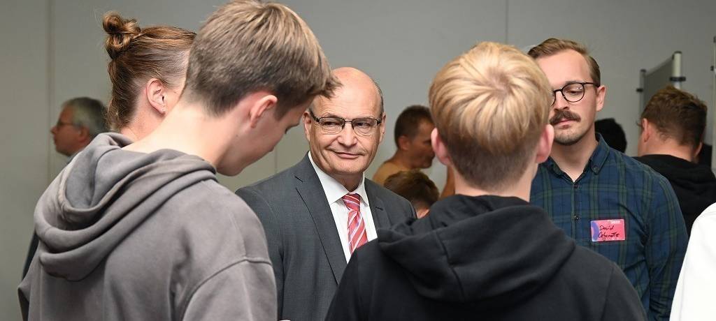 Menschengruppe im Gespräch: Zwei Jugendliche von hinten. Im Zentrum lächelt der Landrat.