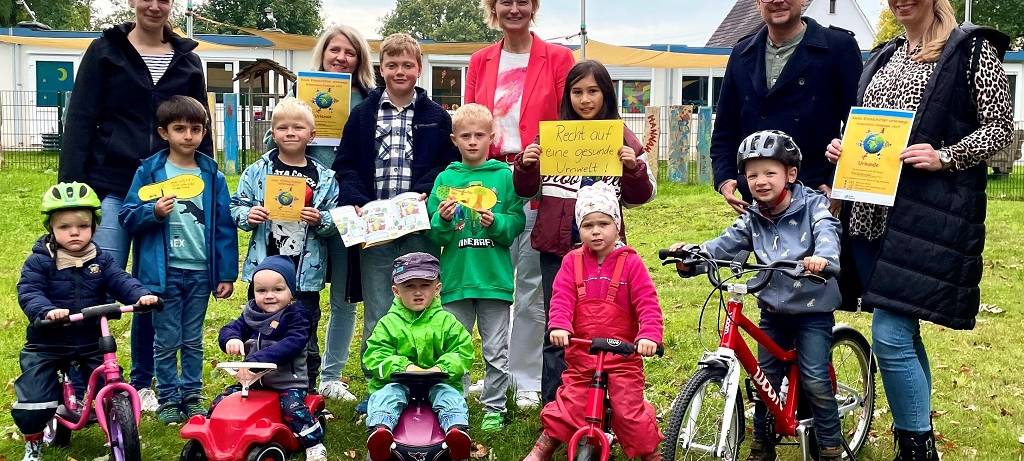 Kindergarten- und Schulkinder in zwei Reihen auf einer Rasenfläche. Vorne die Kleinen mit Bobbycars, Rollern und Fahrrädern, dahinter die Größeren mit Umweltschutz-Transparenten. Ganz hinten Erwachsene.
