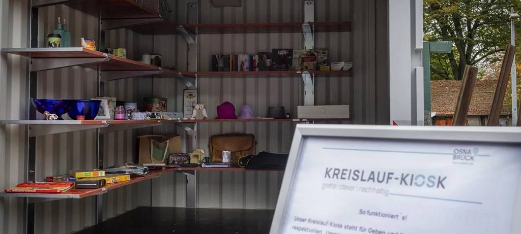 Ein Container mit Regalen voller weiterverwertbarer Dinge. Im Vordergrund ein Aufsteller mit einer Art Gebrauchsanleitung für den Kreislauf-Kiosk.