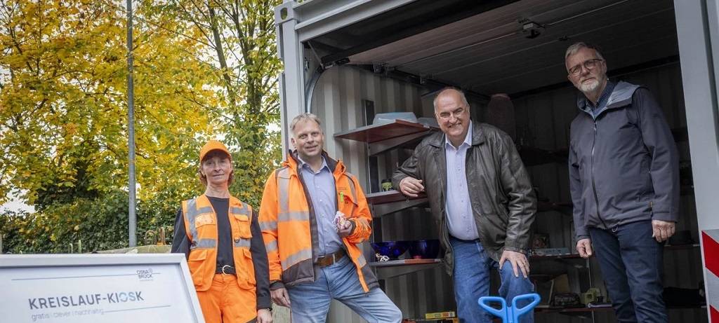 Ein Container mit Regalen voller weiterverwertbarer Dinge. Im Vordergrund eine Frau und ein Mann in orangener Arbeitskleidung und zwei weitere Männer bei der Eröffnung des Kreislauf Kiosk