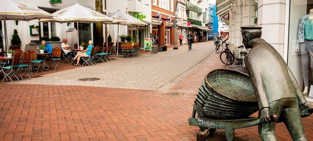 Eine Fußgängerzone mit Außengastronomie und Gästen. Im Vordergrund eine Bronzestatue zur Regionalgeschichte