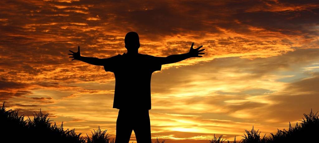 Junger Mann - von hinten aufgenommen, backlighted - breitet die Arme aus vor sonnenrotem Himmel