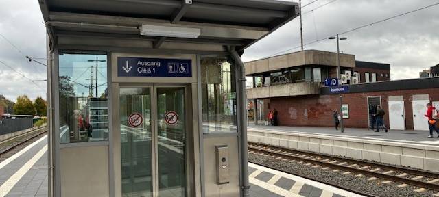 Ein moderner barrierefreier Bahnsteig mit Blindenleitsystem und Aufzug im Vordergrund. Im Hintergrund das ibbenbürener Bahnhofsgebäude