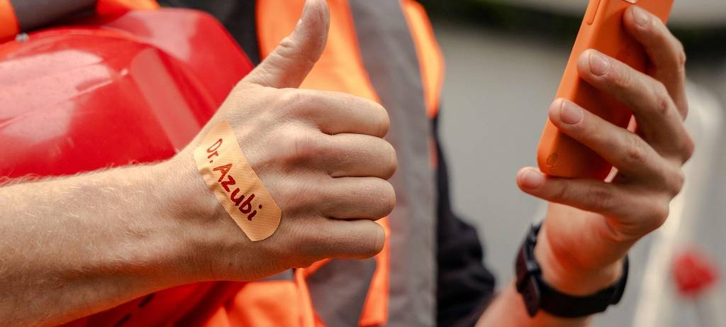 Brustausschnitt eines Arbeiters in orangener Signalkleidung. In der linken Hand ein orangenes Handy, auf dem rechten Handrücken ein orangenes Pflaster mit der Aufschrift "Dr. Azubi"