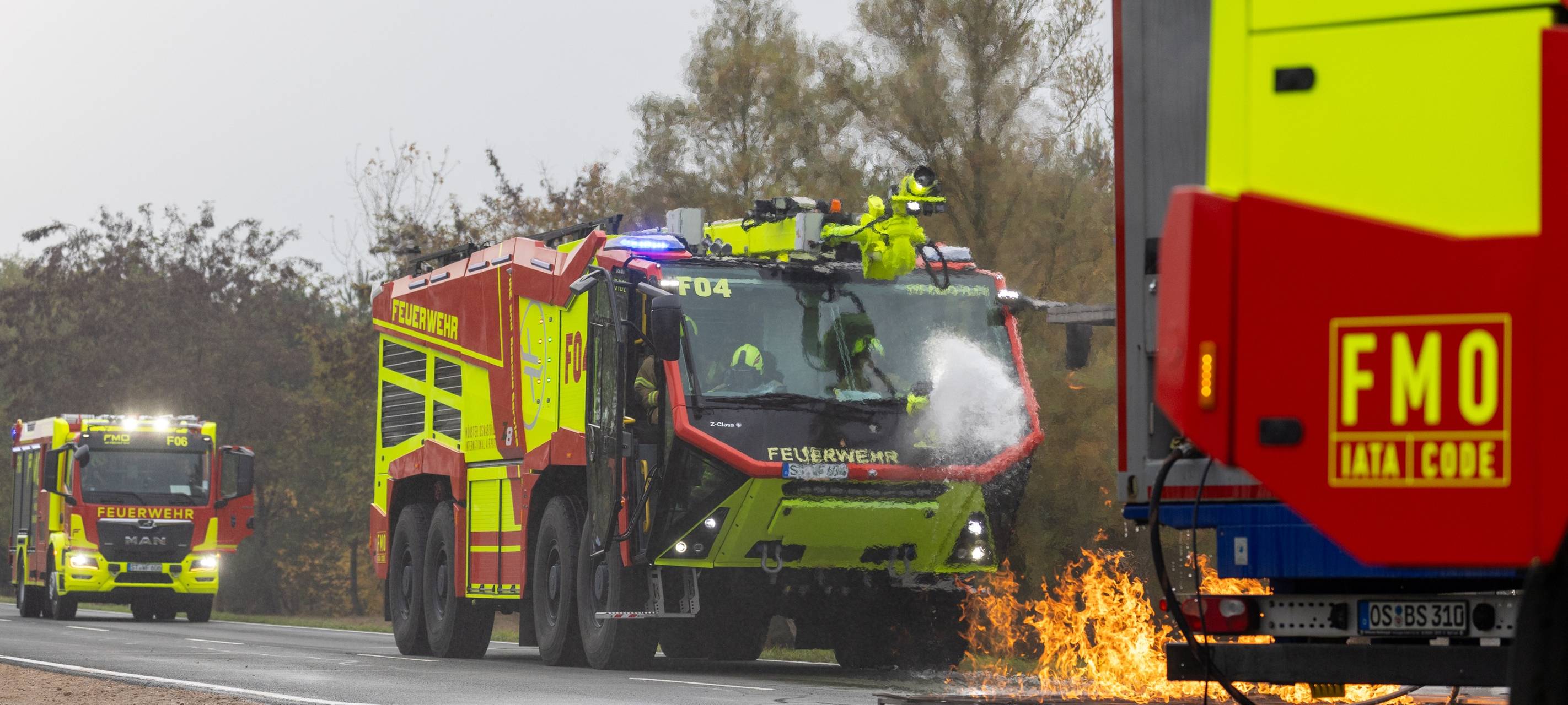 Ein Feuerwehrauto fährt über eine Straße am Flughafen Münster/Osnabrück. Vor dem Auto brennt es.