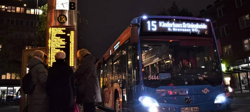 Ein Linienbus im Dunklen und Menschen, die an der Bushaltestelle stehen. Im Hintergrund ein digitaler Fahrplan.