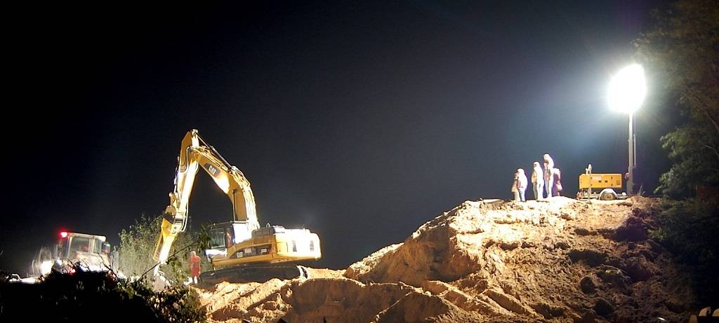 Ein Bagger, der nachts bei künstlicher Beleuchtung auf einer Baustelle arbeitet. Am rechten Rand des Bildes einige Schaulustige