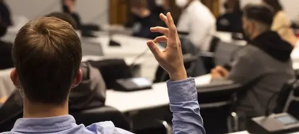 Ein Sitzungssaal mit jungen Menschen. Im Vordergrund ein junger Mann von hinten, der die rechte Hand hebt. Die anderen im Hintergrund unscharf.