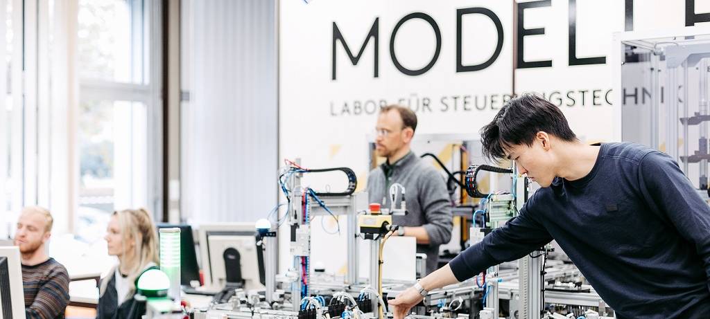Studentinnen und Studenten um eine technische Versuchs-Anordnung. Rechts im Vordergrund legt ein asiatisch aussehender Student Hand an, im Hintergrund der Dozent, der verfolgt, was eine Studentin und ihr Kommilitone auf dem Bildschirm bearbeiten.