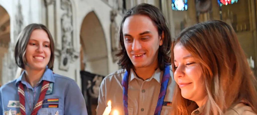 Zwei Pfadfinderinnen und ein Pfadfinder in Uniform mit Kerzen in der Hand beim Empfang des Friedenslichts im Dom zu Münster. Er zündet seine Kerze an ihrer an. Die zweite Kameradin wartet mit ihrer Kerze. Alle lächeln.