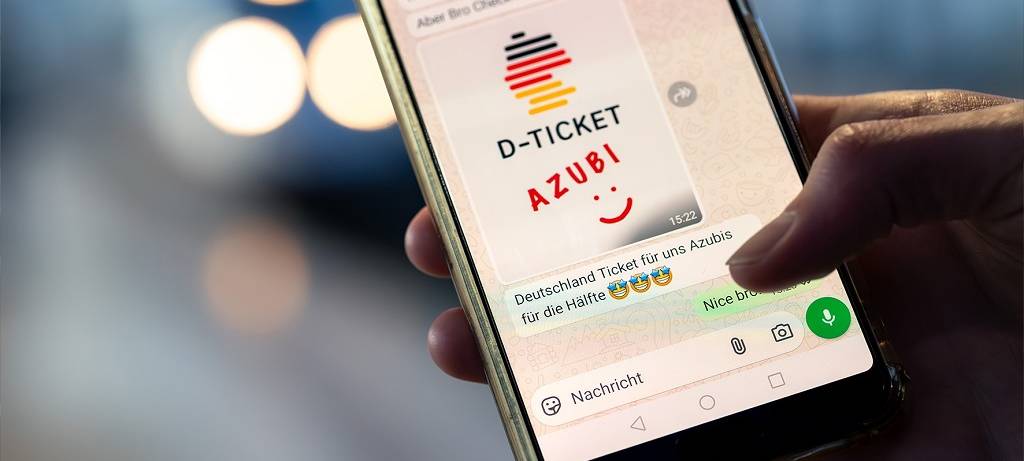 Ein Handy in einer Hand. Auf dem Bildschirm eine WhatsApp-Meldung mit Bild: D-Ticket AZUBI und ein Smiley. Darunter eine Nachricht: Deutschland Ticket für uns Azubis für die Hälfte. Und drei Strahle-Smileys
