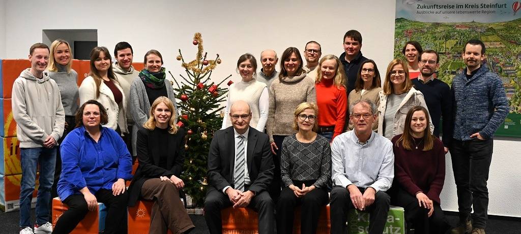 22 lächelnde Offizielle um einen Weihnachtsbaum. Vier von ihnen sitzen im Vordergrund, die anderen stehen dahinter