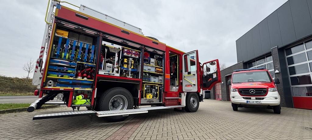 Zwei Feuerwehrfahrzeuge - eins davon offen und voller Technik - auf einem gepflasterten Platz vor einer Halle mit Toren.
